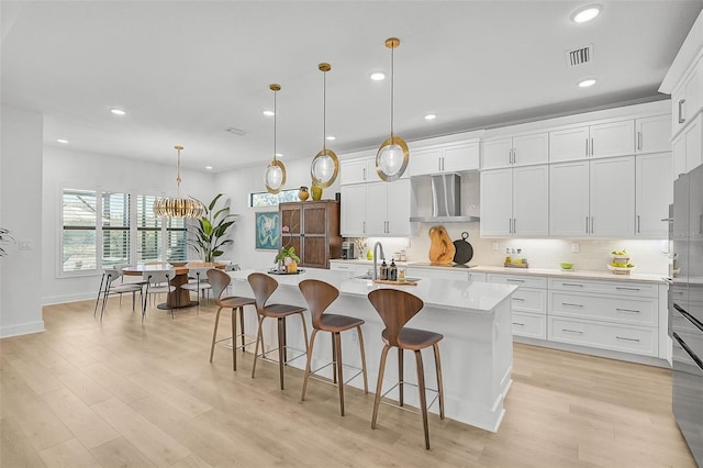 kitchen featuring white cabinetry, wall chimney range hood, pendant lighting, a kitchen island with sink, and a breakfast bar