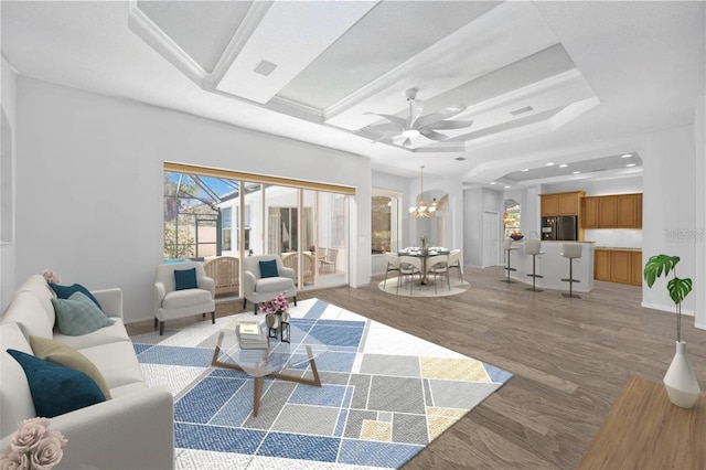 living room featuring light wood-type flooring, ceiling fan with notable chandelier, and a tray ceiling