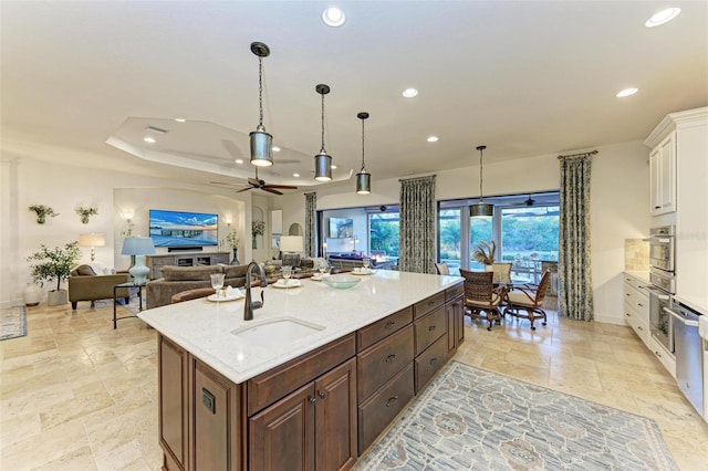 kitchen with light stone counters, a tray ceiling, sink, a center island with sink, and white cabinets