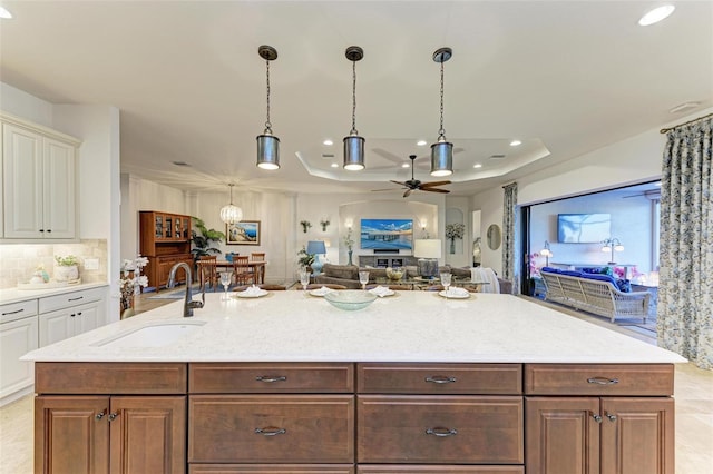 kitchen with light stone counters, a raised ceiling, ceiling fan, sink, and an island with sink