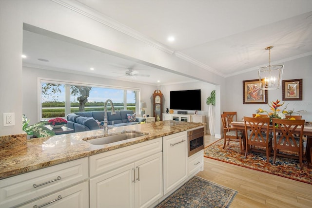 kitchen with light stone counters, ornamental molding, ceiling fan with notable chandelier, sink, and white cabinetry