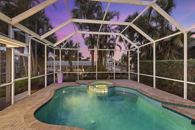 pool at dusk featuring an in ground hot tub, a water view, glass enclosure, and a patio area