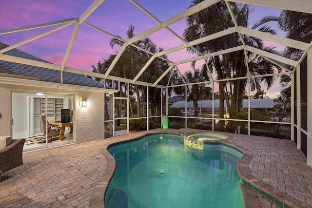 pool at dusk featuring glass enclosure, an in ground hot tub, and a patio