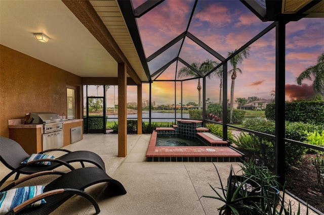 patio terrace at dusk featuring grilling area, glass enclosure, an outdoor kitchen, and an in ground hot tub