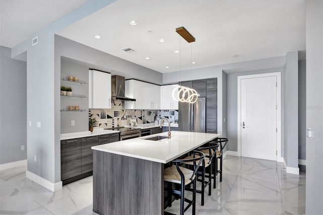 kitchen featuring sink, wall chimney range hood, decorative light fixtures, a kitchen island with sink, and white cabinets