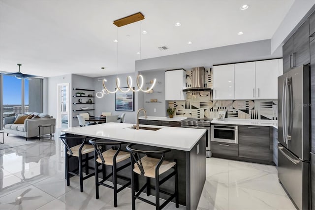 kitchen featuring a center island with sink, wall chimney range hood, sink, hanging light fixtures, and appliances with stainless steel finishes