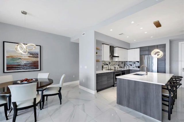 kitchen featuring wall chimney exhaust hood, stainless steel appliances, hanging light fixtures, a kitchen island with sink, and white cabinets