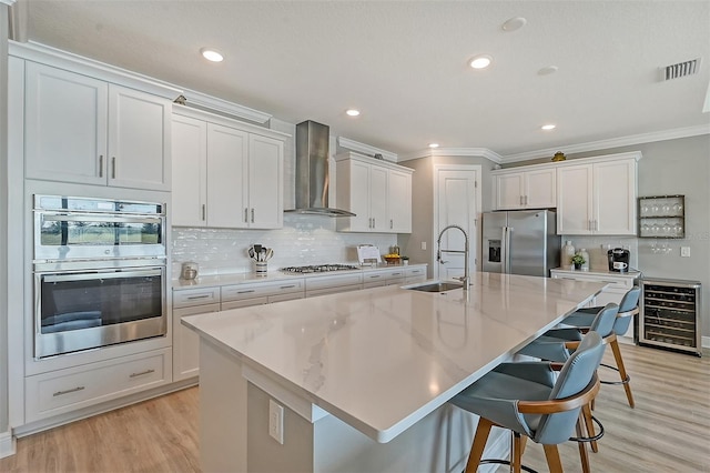 kitchen with sink, beverage cooler, wall chimney range hood, an island with sink, and appliances with stainless steel finishes