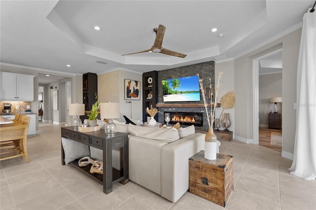 tiled living room with a large fireplace, a raised ceiling, ceiling fan, and ornamental molding