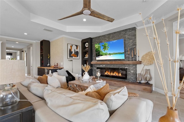 living room featuring a tray ceiling, a stone fireplace, ceiling fan, and ornamental molding