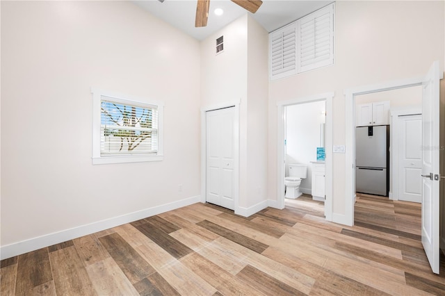 unfurnished bedroom with ensuite bathroom, a towering ceiling, ceiling fan, light wood-type flooring, and stainless steel fridge