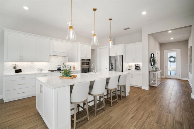 kitchen featuring white cabinetry, backsplash, a center island with sink, appliances with stainless steel finishes, and custom exhaust hood