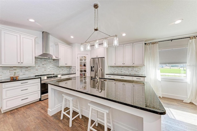 kitchen featuring a large island with sink, wall chimney range hood, vaulted ceiling, appliances with stainless steel finishes, and white cabinetry