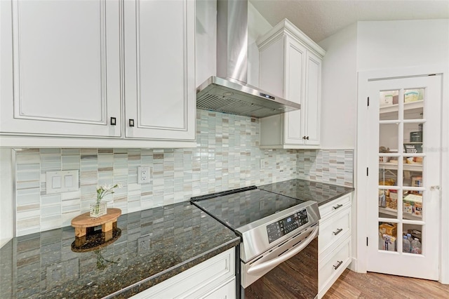 kitchen with wall chimney range hood, backsplash, dark stone counters, stainless steel electric stove, and white cabinets