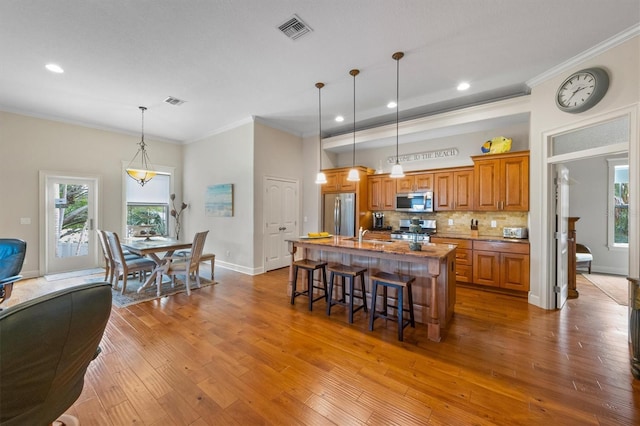 kitchen with a kitchen island with sink, stainless steel appliances, hanging light fixtures, crown molding, and a breakfast bar