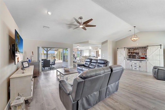 living room with ceiling fan with notable chandelier, light wood-type flooring, vaulted ceiling, and a barn door