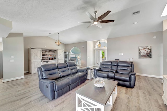 living room with ceiling fan, a barn door, a textured ceiling, vaulted ceiling, and light wood-type flooring