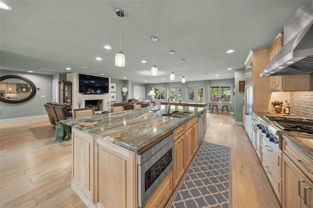 kitchen featuring a center island with sink, wall chimney range hood, sink, dark stone countertops, and stainless steel appliances