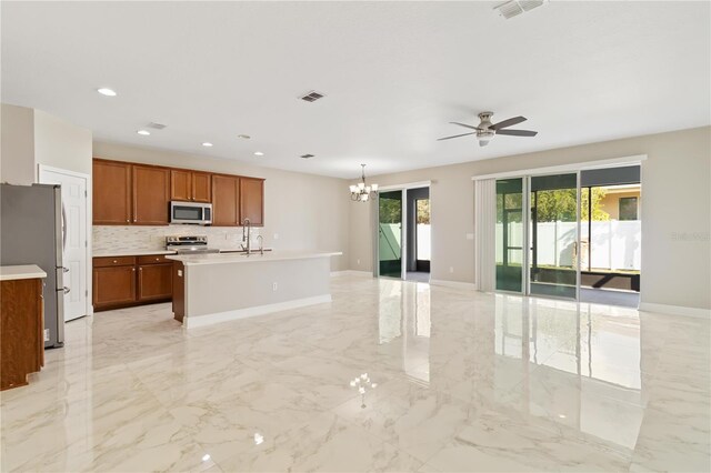 kitchen with tasteful backsplash, ceiling fan with notable chandelier, stainless steel appliances, pendant lighting, and an island with sink