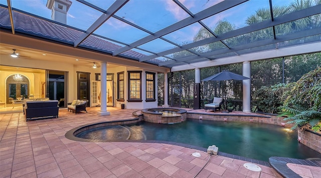 pool at dusk featuring glass enclosure, ceiling fan, pool water feature, a patio area, and an in ground hot tub