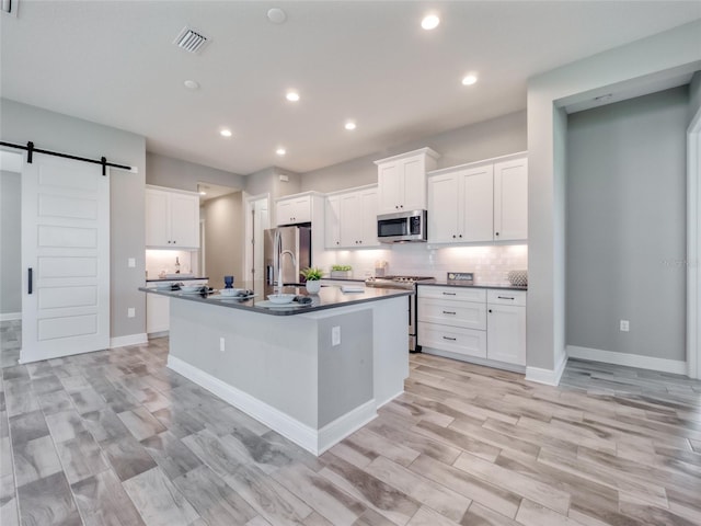 kitchen with a barn door, white cabinetry, appliances with stainless steel finishes, dark countertops, and a center island with sink