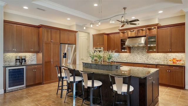 kitchen featuring a kitchen breakfast bar, stainless steel appliances, a kitchen island with sink, beverage cooler, and dark stone countertops