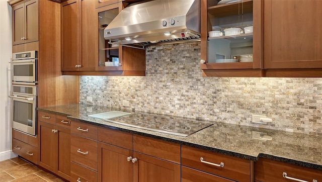 kitchen with wall chimney exhaust hood, backsplash, dark stone counters, light tile patterned flooring, and electric stovetop