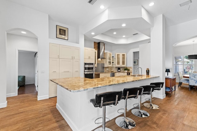 kitchen with kitchen peninsula, wall chimney exhaust hood, a breakfast bar, stainless steel appliances, and sink