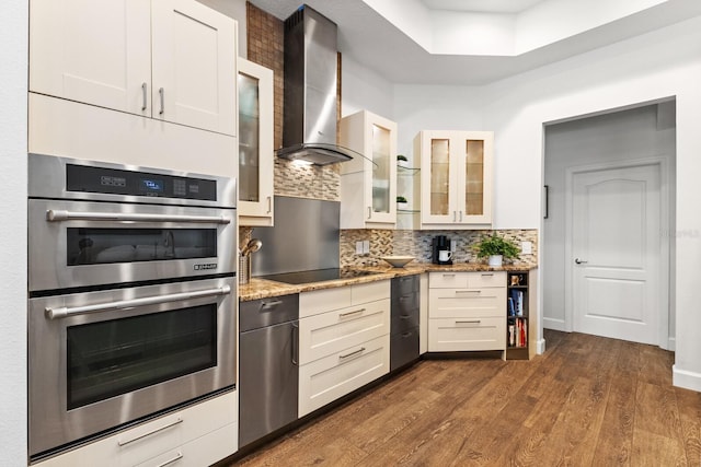 kitchen featuring light stone countertops, black electric stovetop, double oven, wall chimney range hood, and white cabinetry