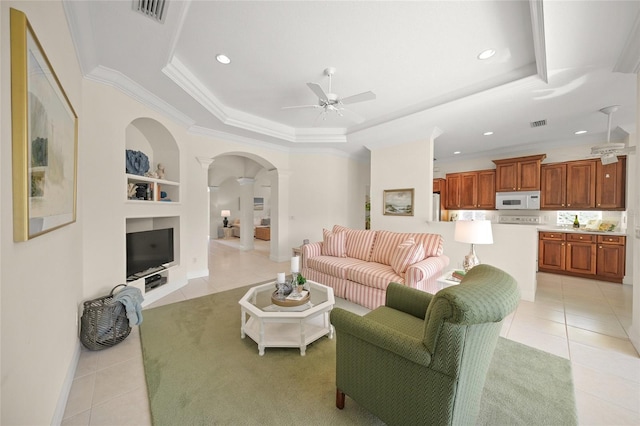 tiled living room with ornate columns, built in shelves, a tray ceiling, ceiling fan, and crown molding