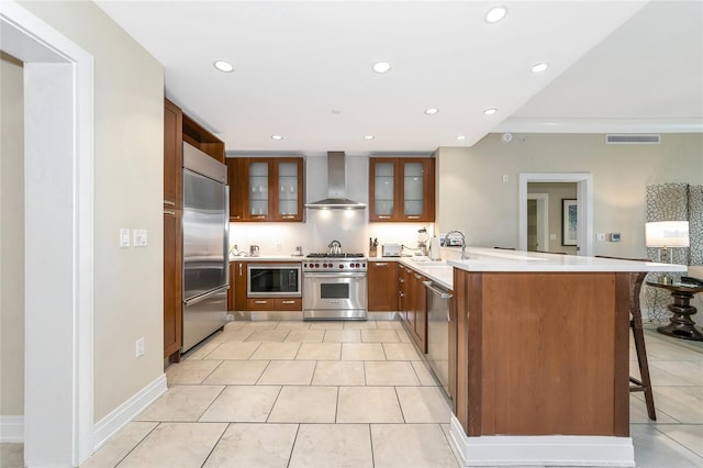 kitchen with a kitchen breakfast bar, built in appliances, wall chimney exhaust hood, light tile patterned floors, and kitchen peninsula