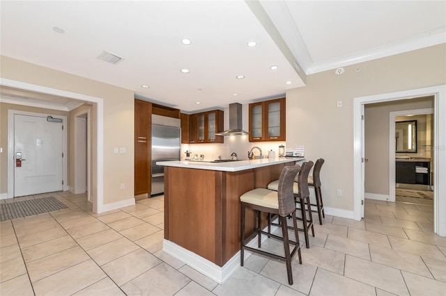 kitchen with a breakfast bar, stainless steel built in fridge, wall chimney range hood, crown molding, and kitchen peninsula