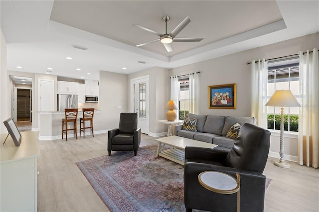 living room with light wood-type flooring, a tray ceiling, a wealth of natural light, and ceiling fan