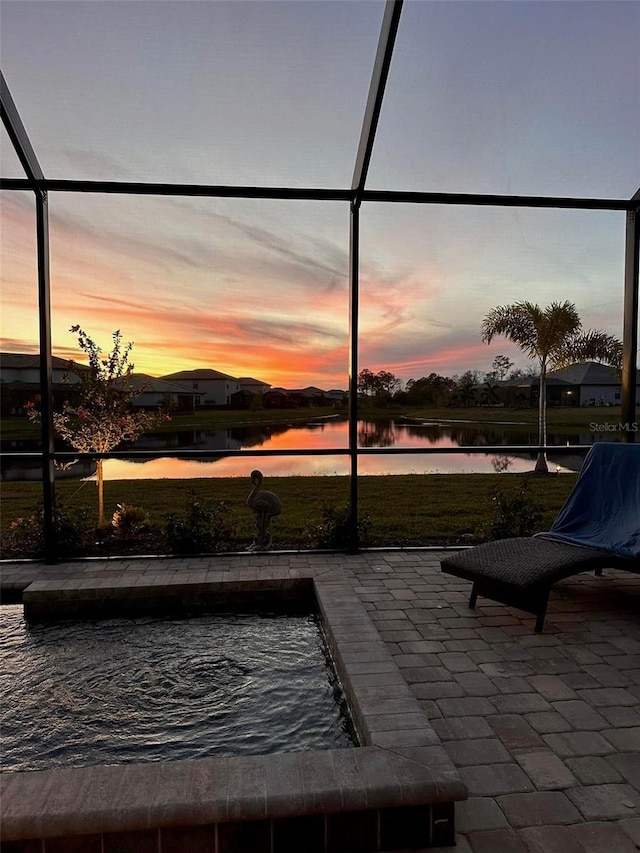 pool at dusk with glass enclosure, a water view, and a patio