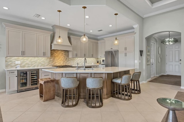 kitchen with stainless steel appliances, decorative light fixtures, custom range hood, an island with sink, and wine cooler