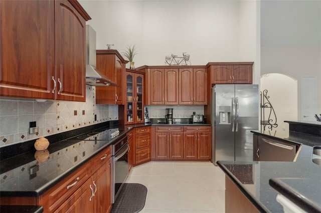 kitchen featuring light tile patterned flooring, appliances with stainless steel finishes, a towering ceiling, decorative backsplash, and wall chimney range hood