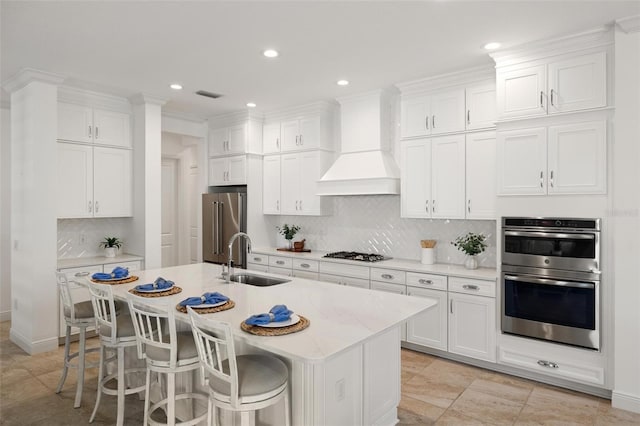 kitchen with appliances with stainless steel finishes, premium range hood, sink, white cabinetry, and an island with sink