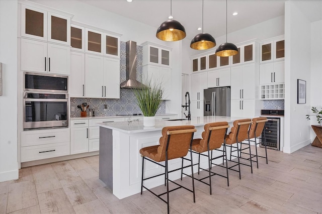 kitchen featuring appliances with stainless steel finishes, wall chimney range hood, sink, decorative light fixtures, and an island with sink