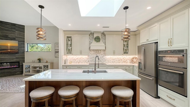 kitchen with appliances with stainless steel finishes, a kitchen island with sink, a skylight, light stone counters, and custom exhaust hood