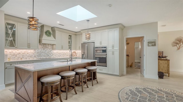 kitchen featuring premium range hood, a skylight, decorative backsplash, hanging light fixtures, and a center island with sink