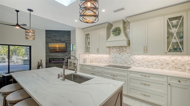 kitchen with premium range hood, sink, decorative light fixtures, an island with sink, and black electric stovetop