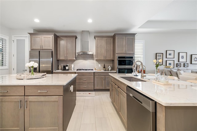 kitchen with sink, wall chimney exhaust hood, light stone countertops, an island with sink, and stainless steel appliances