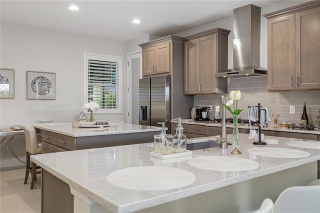 kitchen with decorative backsplash, light stone counters, wall chimney range hood, stainless steel fridge with ice dispenser, and an island with sink