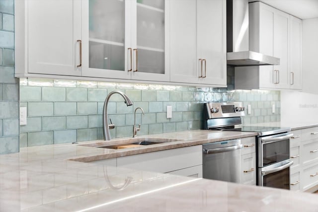 kitchen with sink, wall chimney exhaust hood, backsplash, white cabinets, and appliances with stainless steel finishes