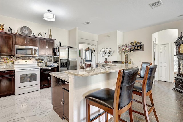 kitchen featuring decorative backsplash, light stone countertops, dark brown cabinetry, stainless steel appliances, and a breakfast bar area