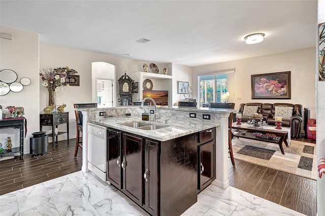 kitchen featuring dishwasher, a center island with sink, sink, light stone countertops, and dark brown cabinetry