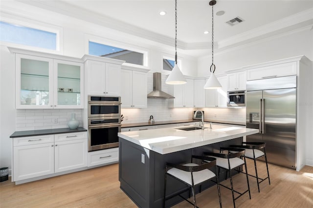 kitchen featuring wall chimney exhaust hood, stainless steel appliances, sink, pendant lighting, and a center island with sink