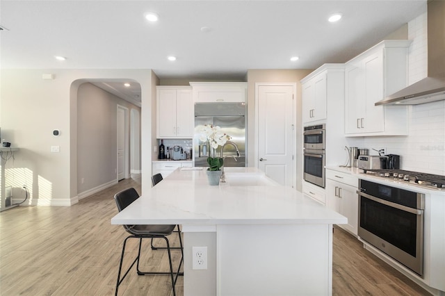 kitchen with appliances with stainless steel finishes, a kitchen breakfast bar, wall chimney exhaust hood, white cabinetry, and an island with sink