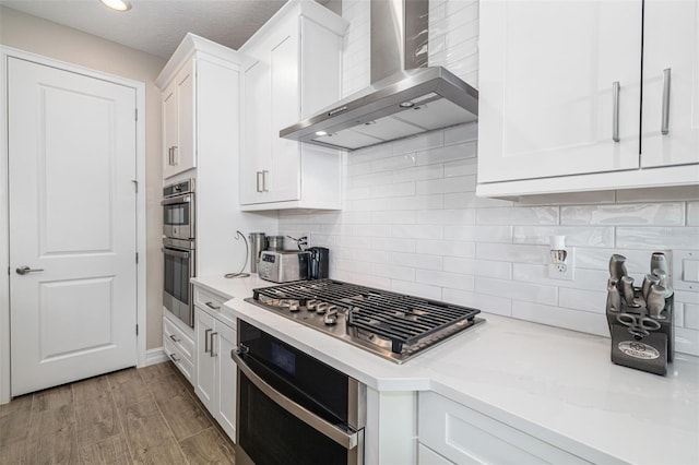 kitchen featuring light stone counters, stainless steel appliances, wall chimney range hood, white cabinets, and light hardwood / wood-style floors