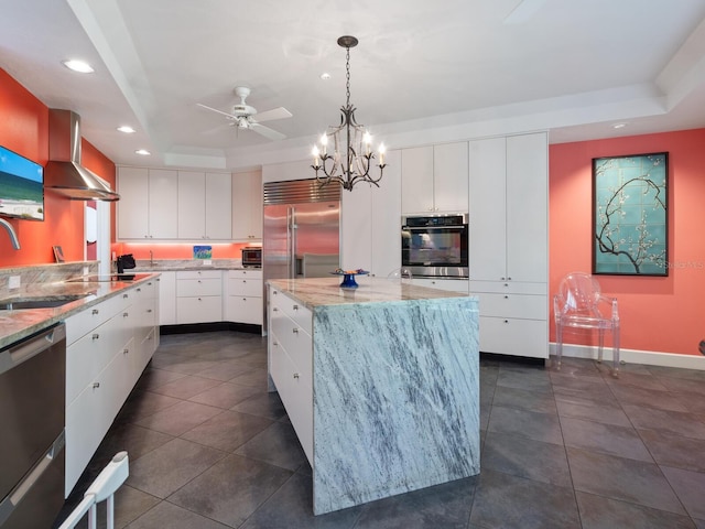 kitchen with a center island, white cabinets, a raised ceiling, wall chimney range hood, and appliances with stainless steel finishes